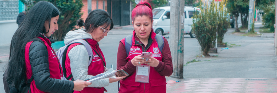 funcionarios de la Secretaría Distrital de Planeación con chaleco Vinotinto, realizando encuesta de Sisbén a un ciudadano sonriente. 