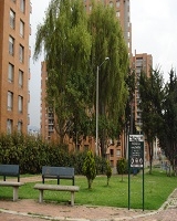 Imagen de una zona verde rodeado de un camino peatonal con sillas para los transeúntes, árboles que colindan con conjuntos de apartamentos.  
