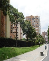 Fotografía De izquierda a derecha se encuentra un conjunto residencial rodeado por cerca viva seguido de árboles altos y delgados luego continua una zona ver alrededor del conjunto, posteriormente un camino peatonal con bancas de descanso y canasta de basura para finalizar con una vía vehicular. 