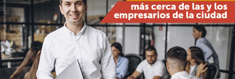 Hombre con portatil en sus manos, al fondo mujeres y hombres reunidos en mesa de trabajo.
